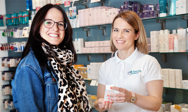 Lächelnde Apothekerin in der Rotenbühl Apotheke in Saarbrücken hält Medikamentenpackung, während sie eine Kundin berät, beides strahlt Freundlichkeit und Professionalität aus.